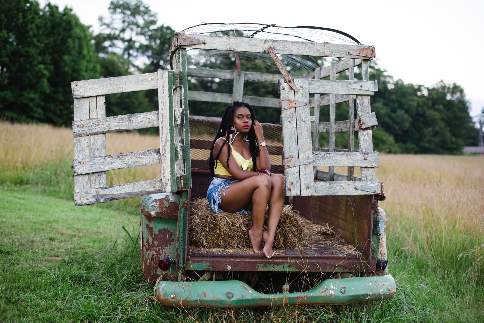 A young woman seated on hay in the back of a weathered old truck in a grassy field, with trees in the background