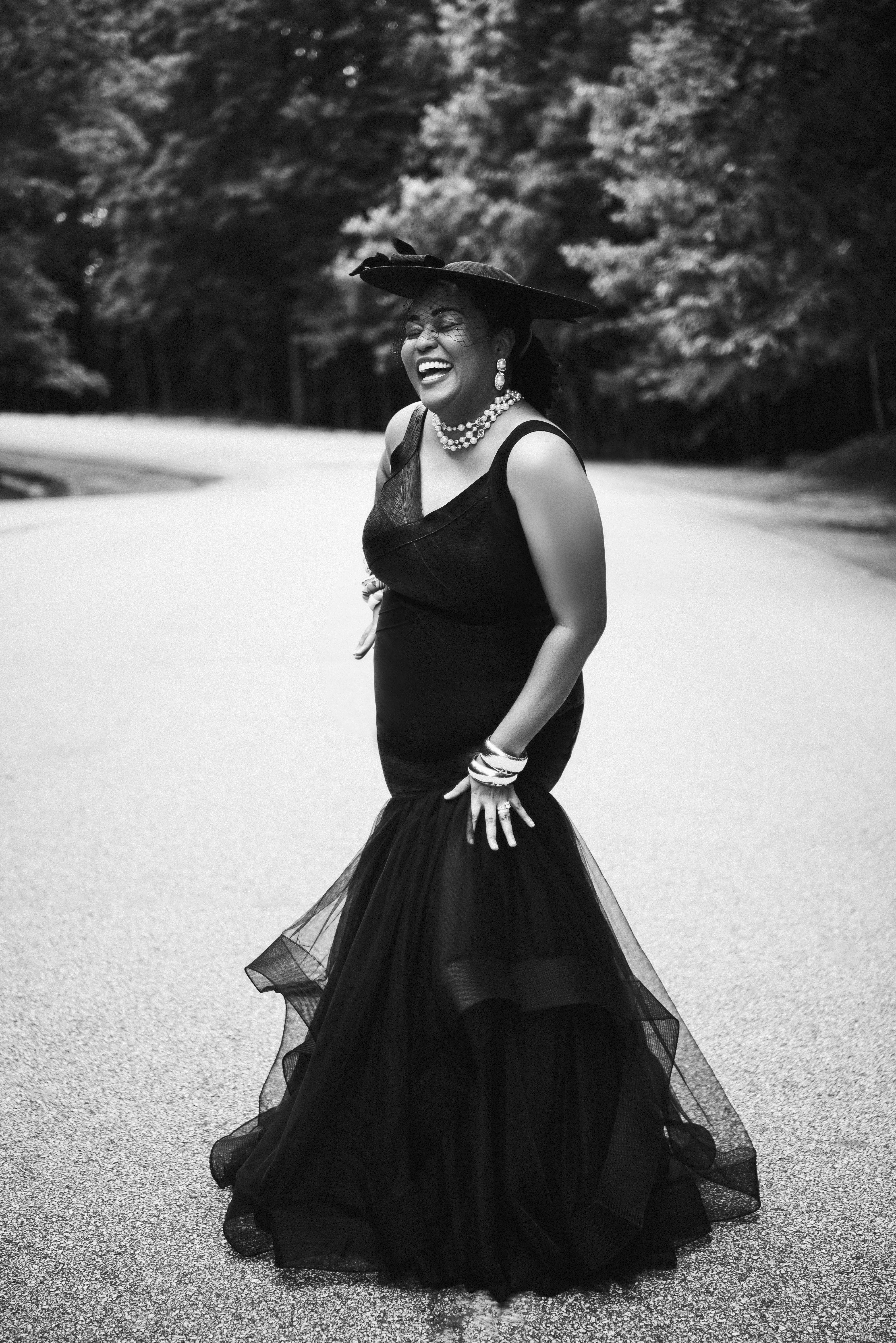 Black and white portrait of a woman in a black dress and veiled hat, laughing on a wooded road