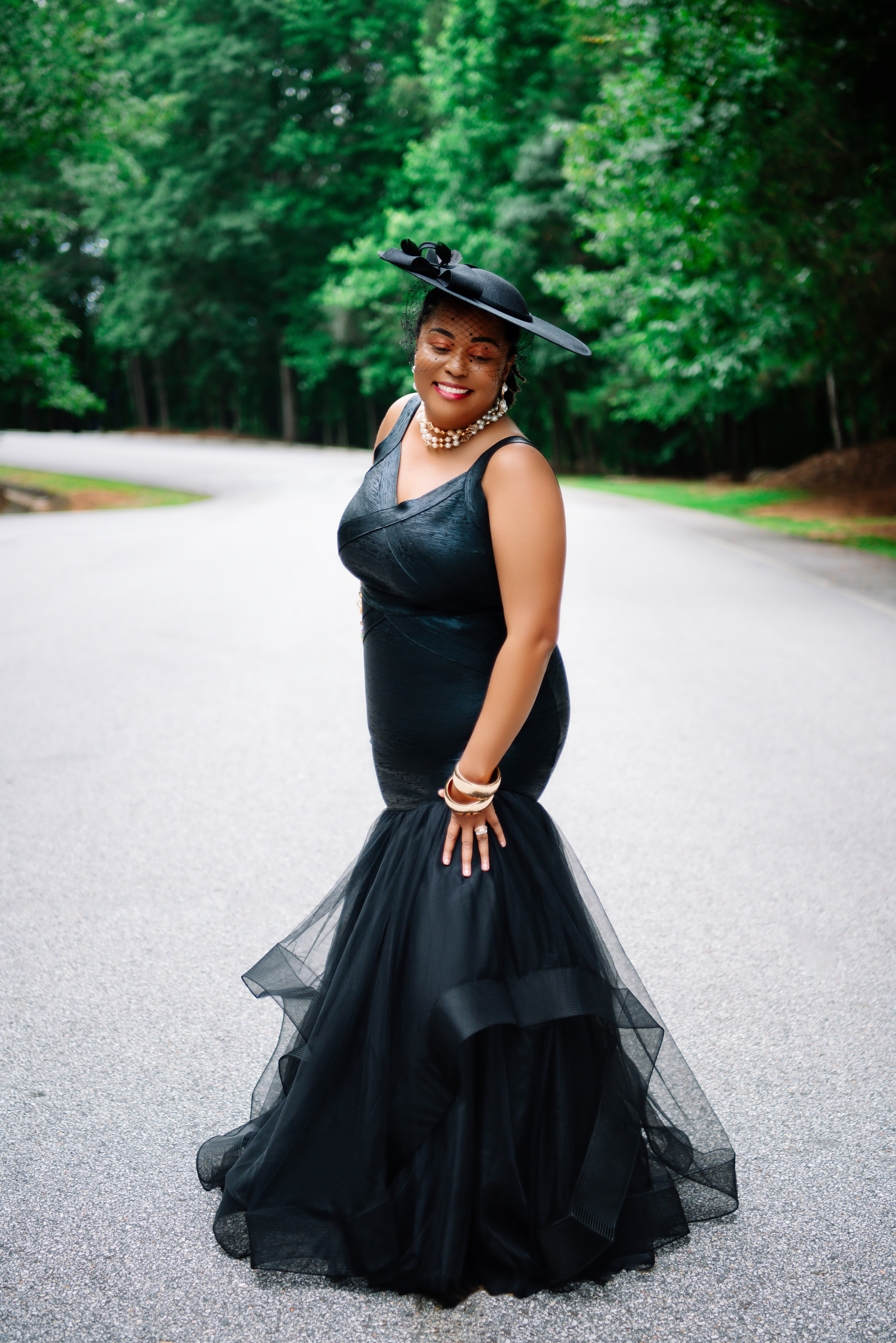 Full-length portrait in a black mermaid-style dress with tulle skirt and a wide-brimmed hat with a veil, smiling on a quiet tree-lined road