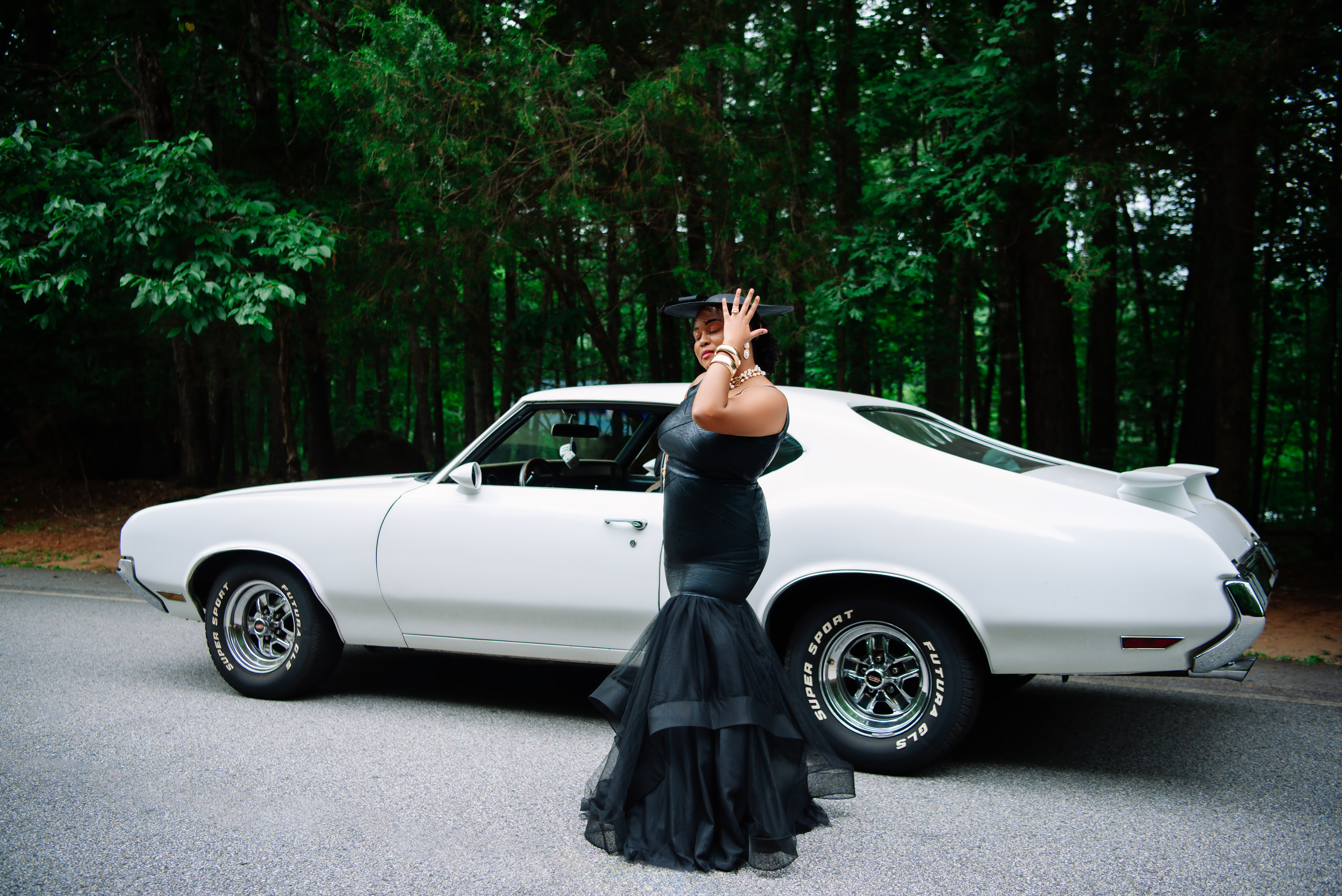 Posing beside a white vintage muscle car in a black dress and veiled hat, hand raised to her hat