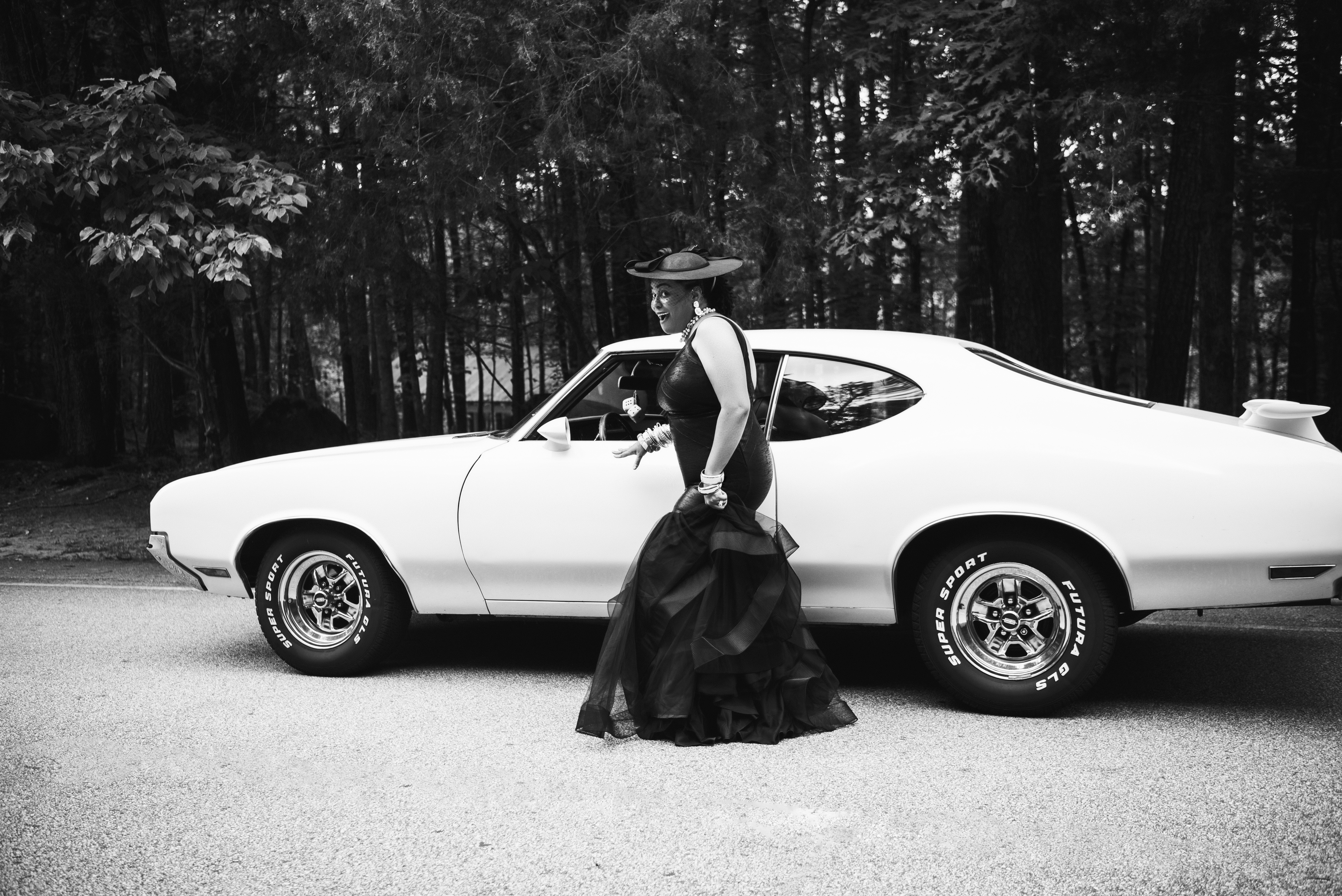 Black and white photograph of her stepping out from beside a vintage muscle car in a black tulle-skirted dress and hat