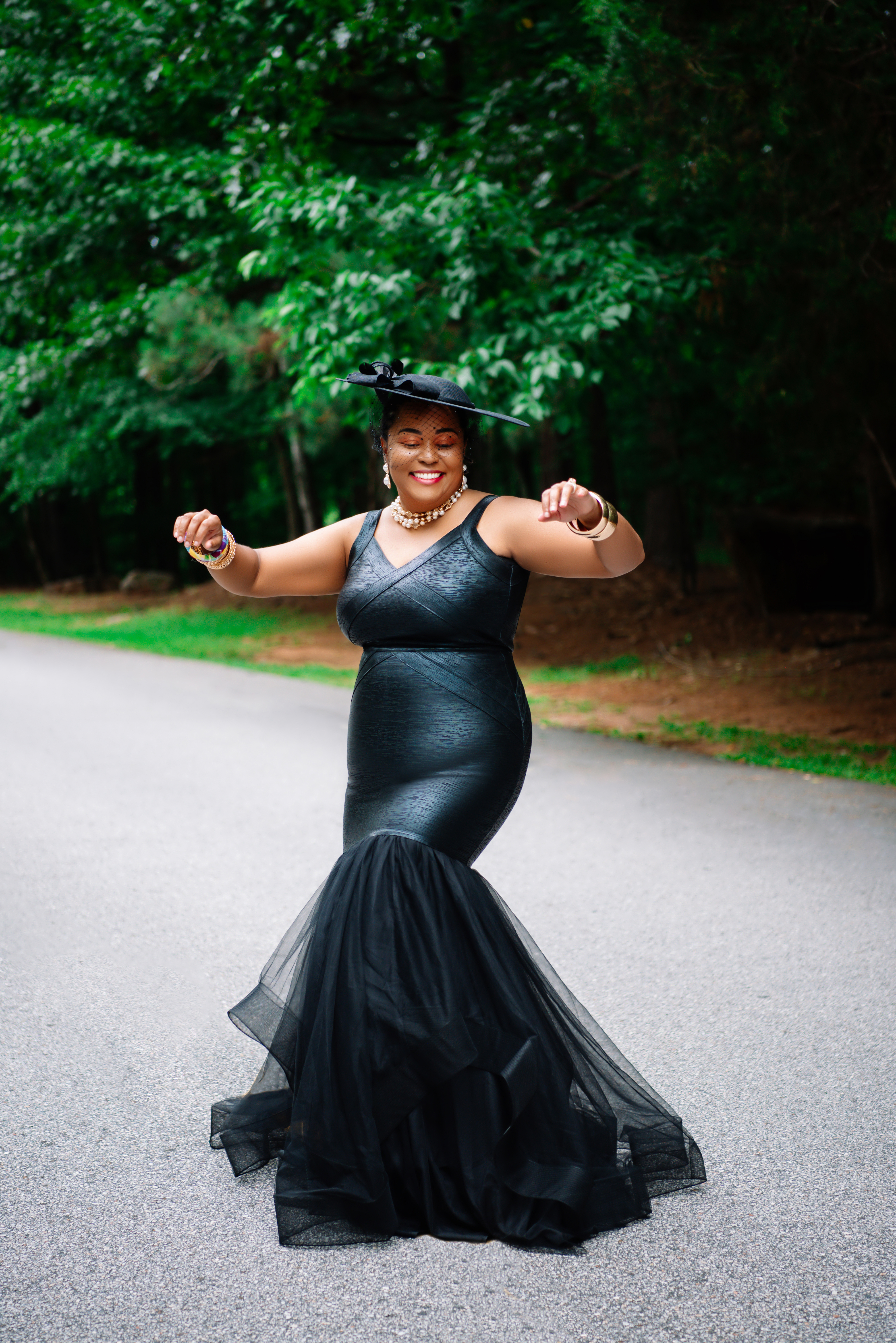 Arms open wide, dancing joyfully in a black dress on a tree-lined road