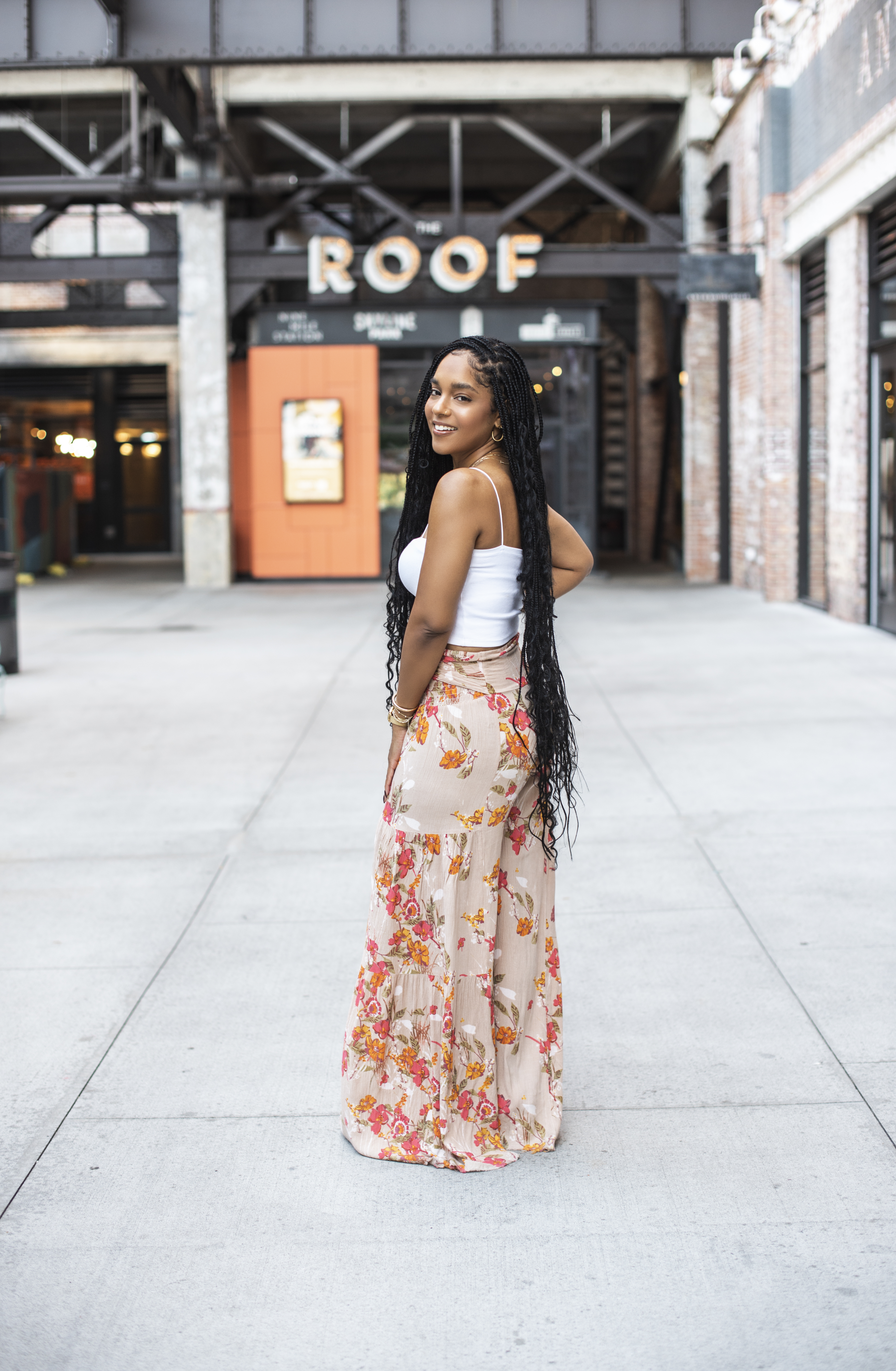 Looking back over her shoulder in floral wide-leg pants and a white tank top in a downtown courtyard