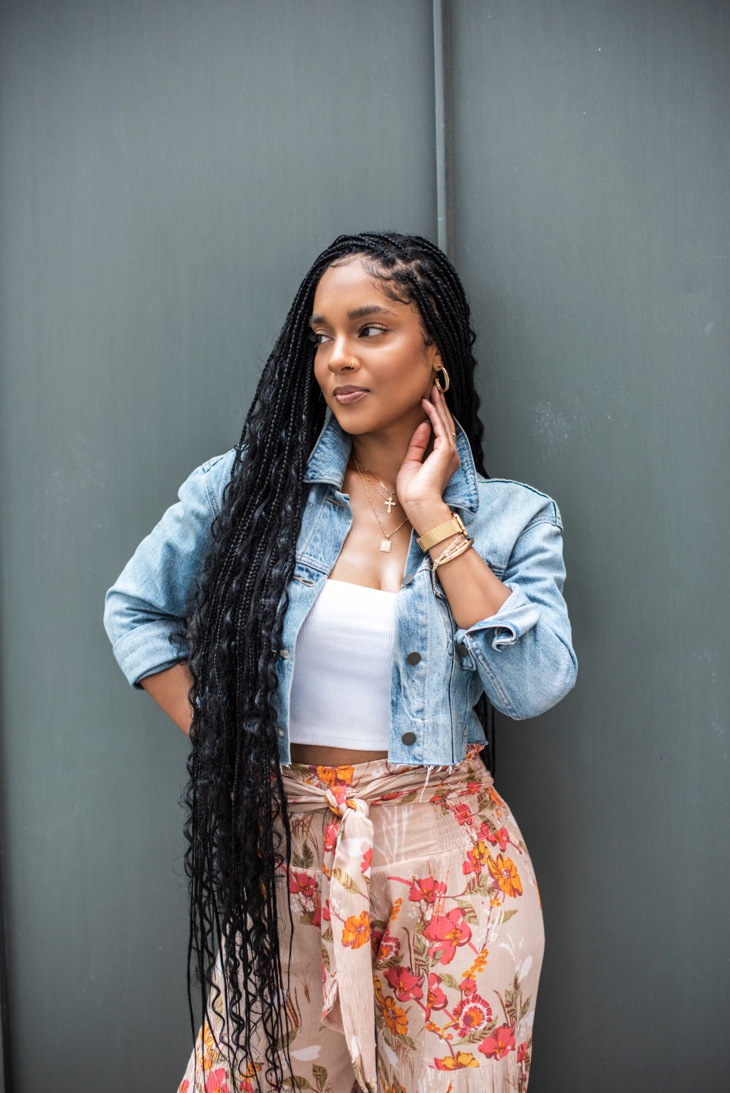 Portrait in a denim jacket and white tank top, hand to her face, long boho braids, against a gray wall