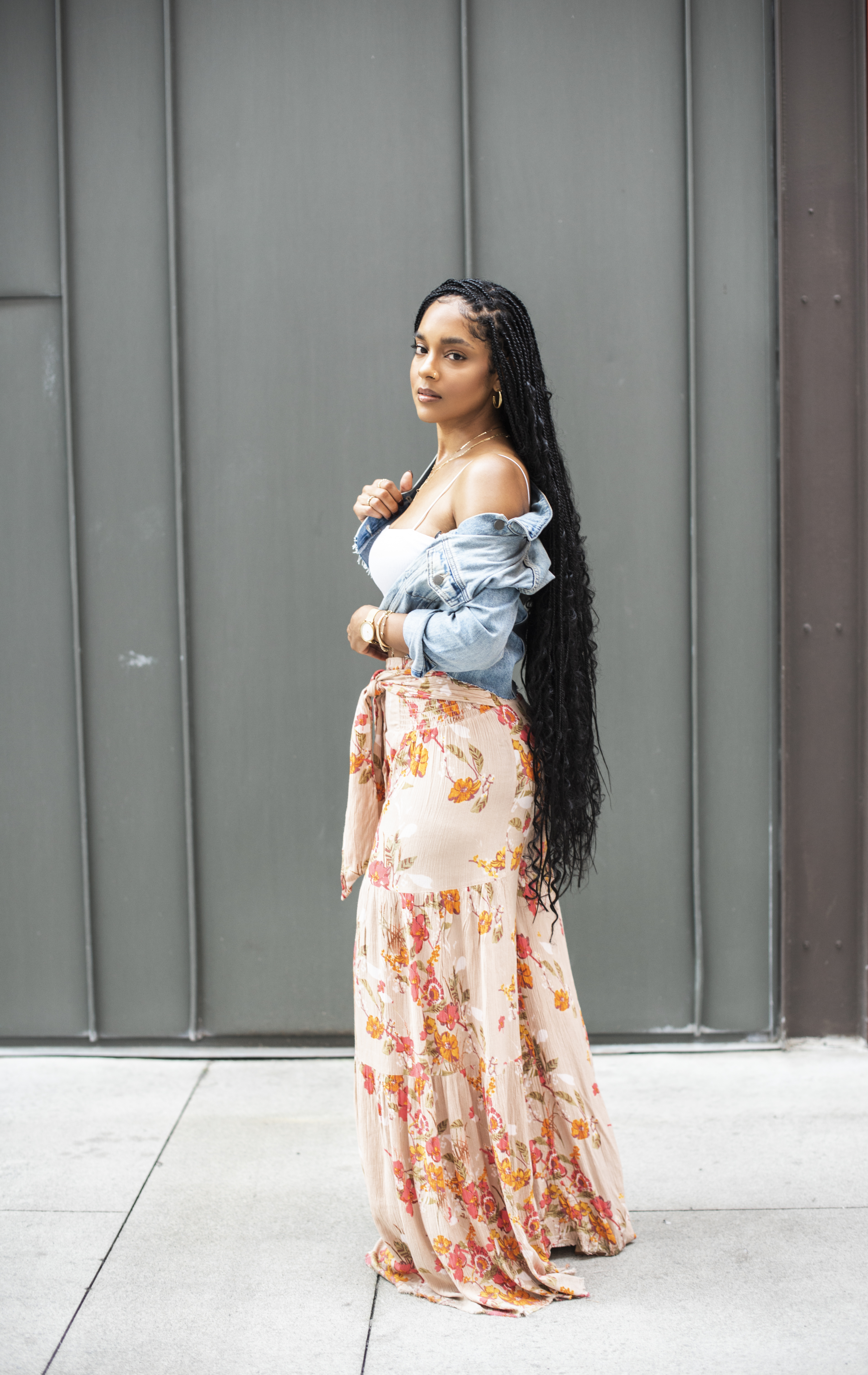 Full-length in floral flowy pants and an off-the-shoulder denim jacket against a gray wall