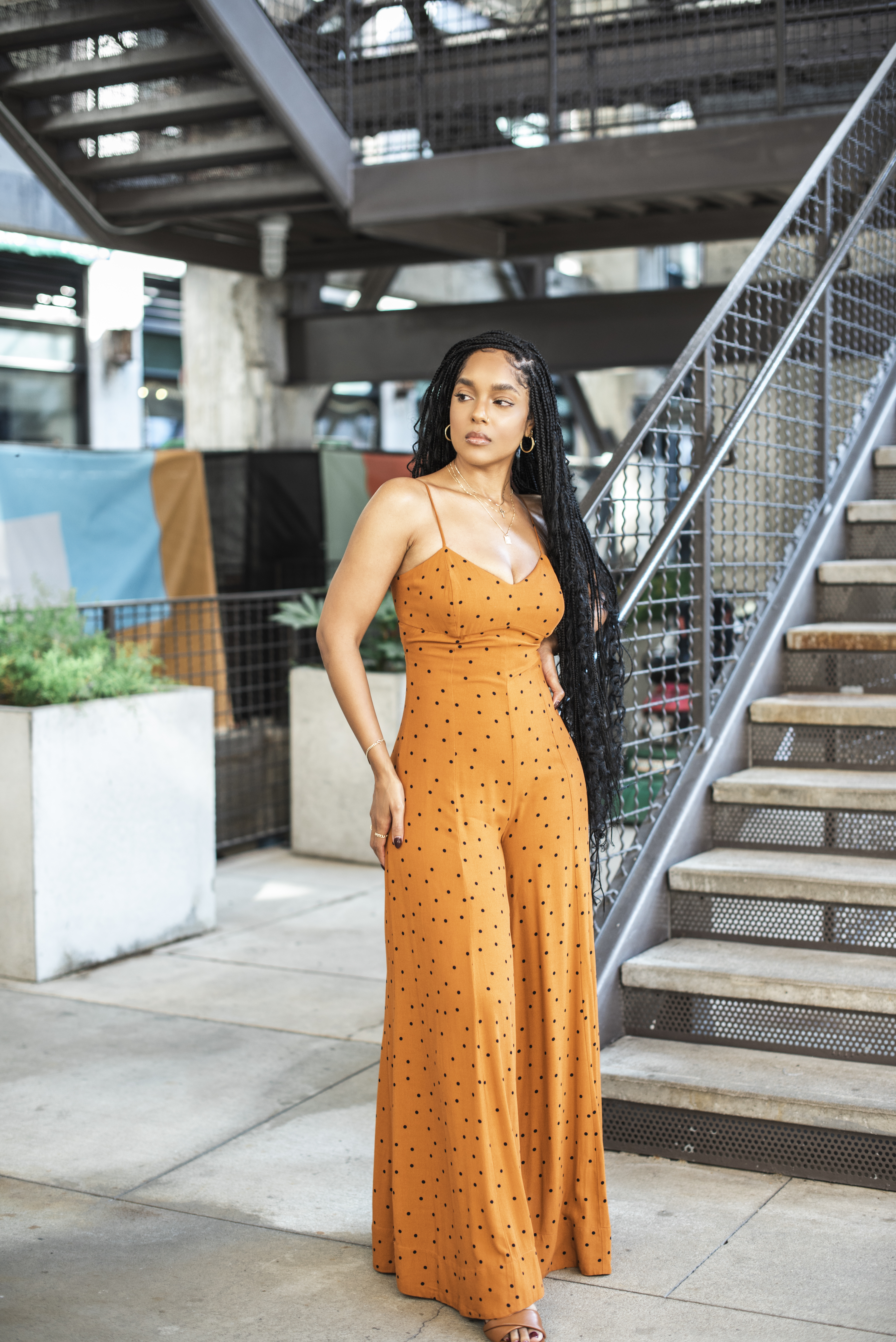 Standing beside an industrial staircase in an orange polka-dot wide-leg jumpsuit