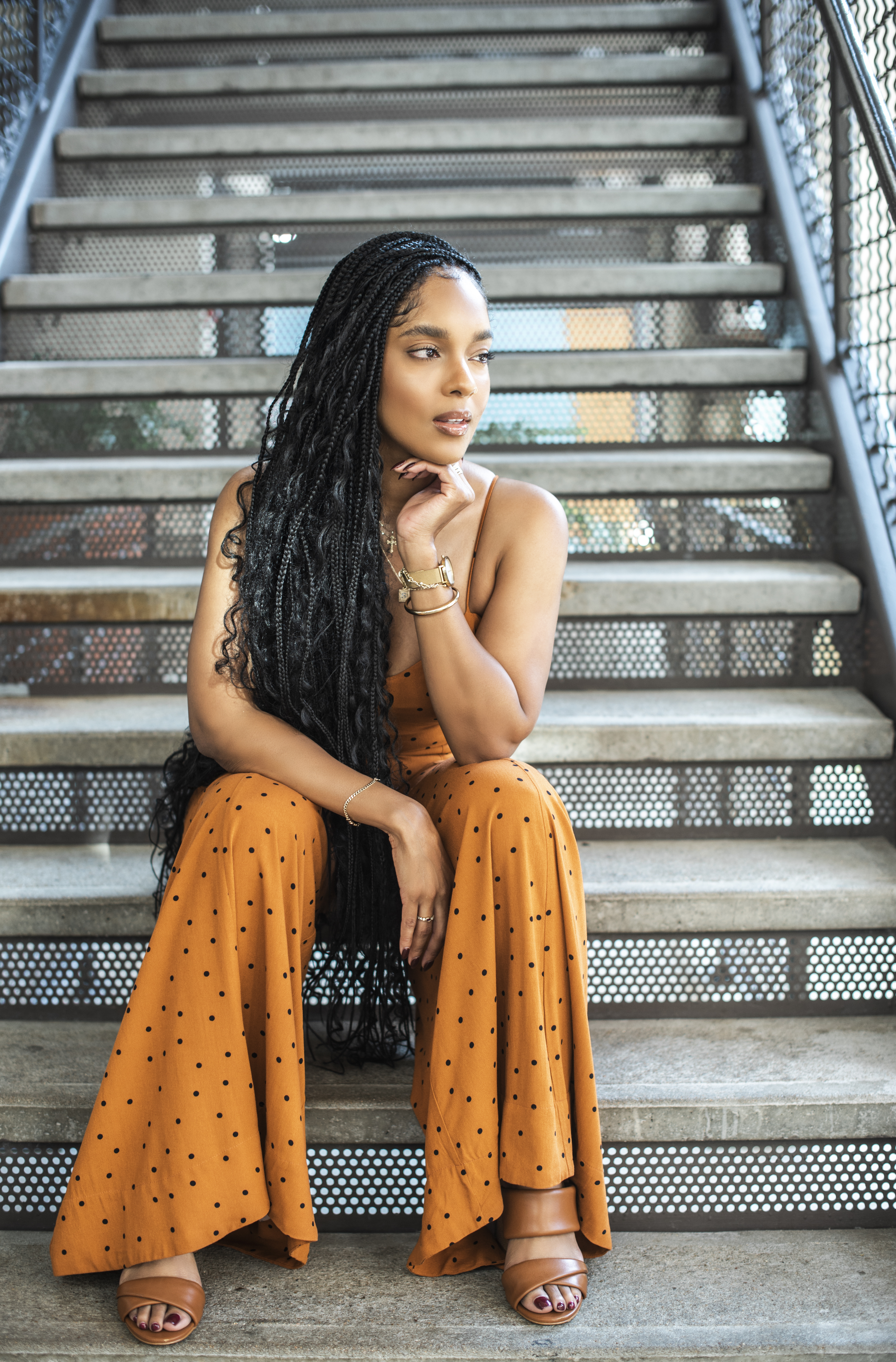 Seated on metal stairs in an orange polka-dot jumpsuit, chin resting on her hand