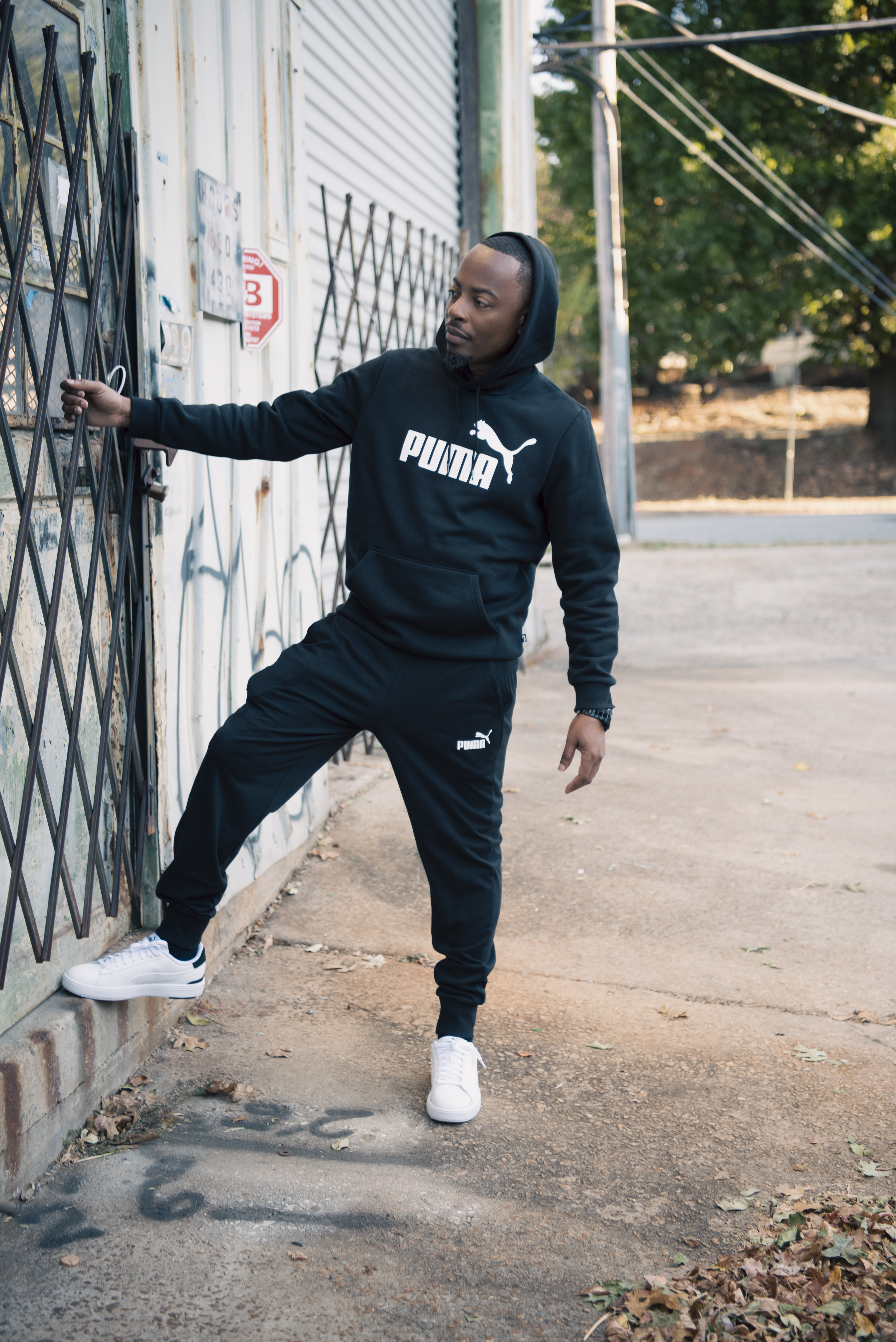 Leaning against a metal gate in a black Puma hoodie, sweatpants and white sneakers