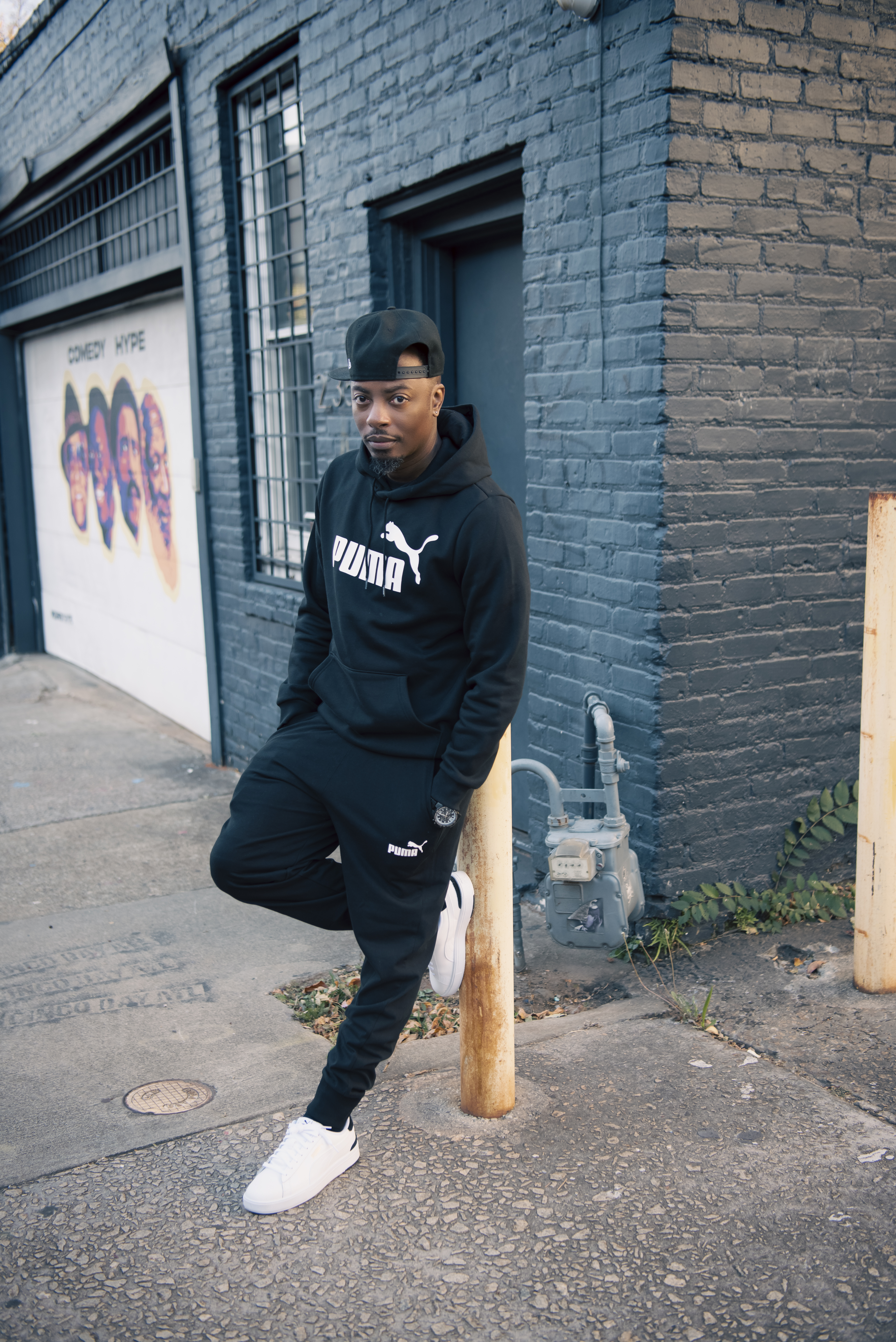 Leaning against a post in a black Puma sweatsuit and white sneakers beside a dark brick building