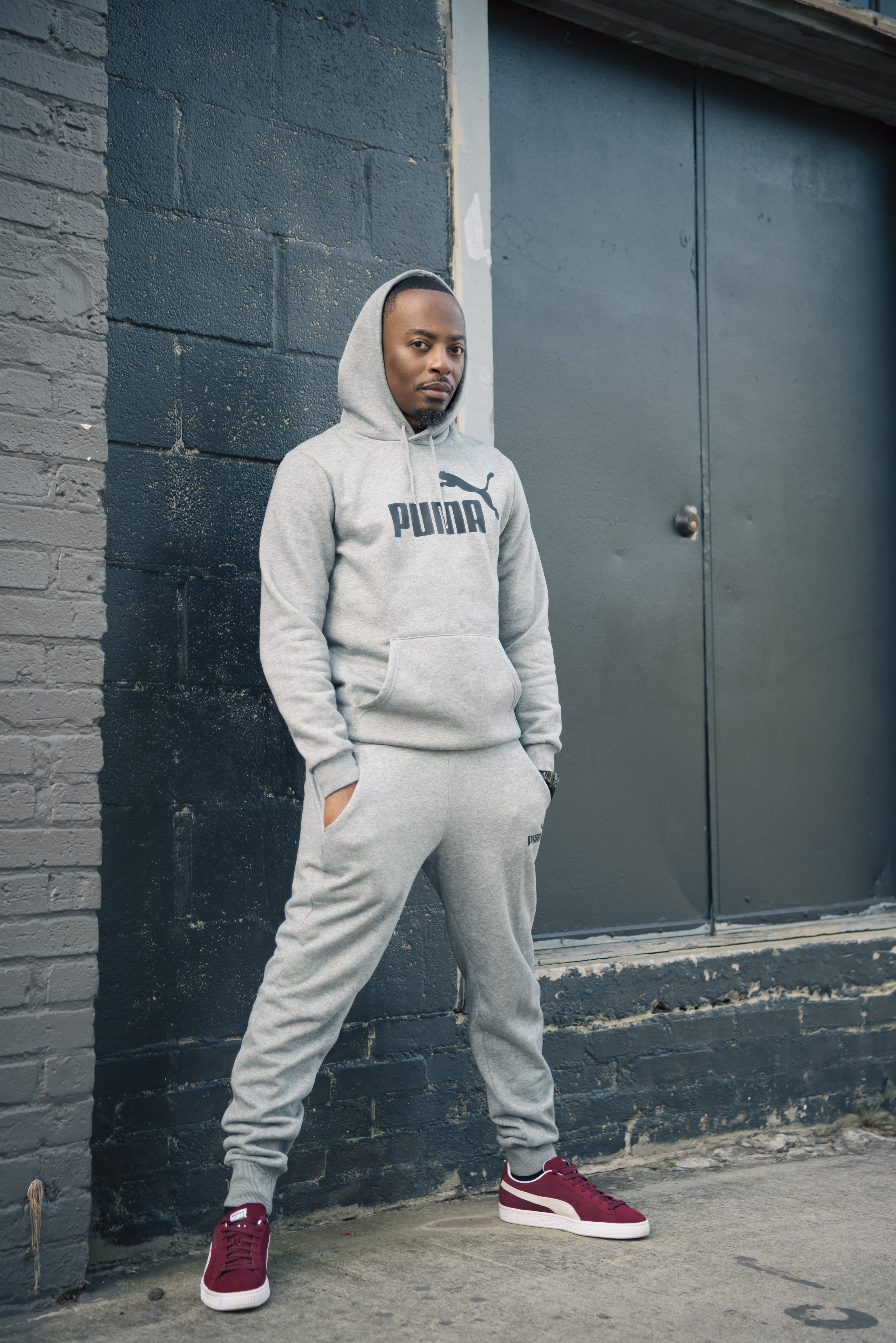 Standing in a gray Puma sweatsuit and burgundy sneakers, hands in pockets, in front of a metal door