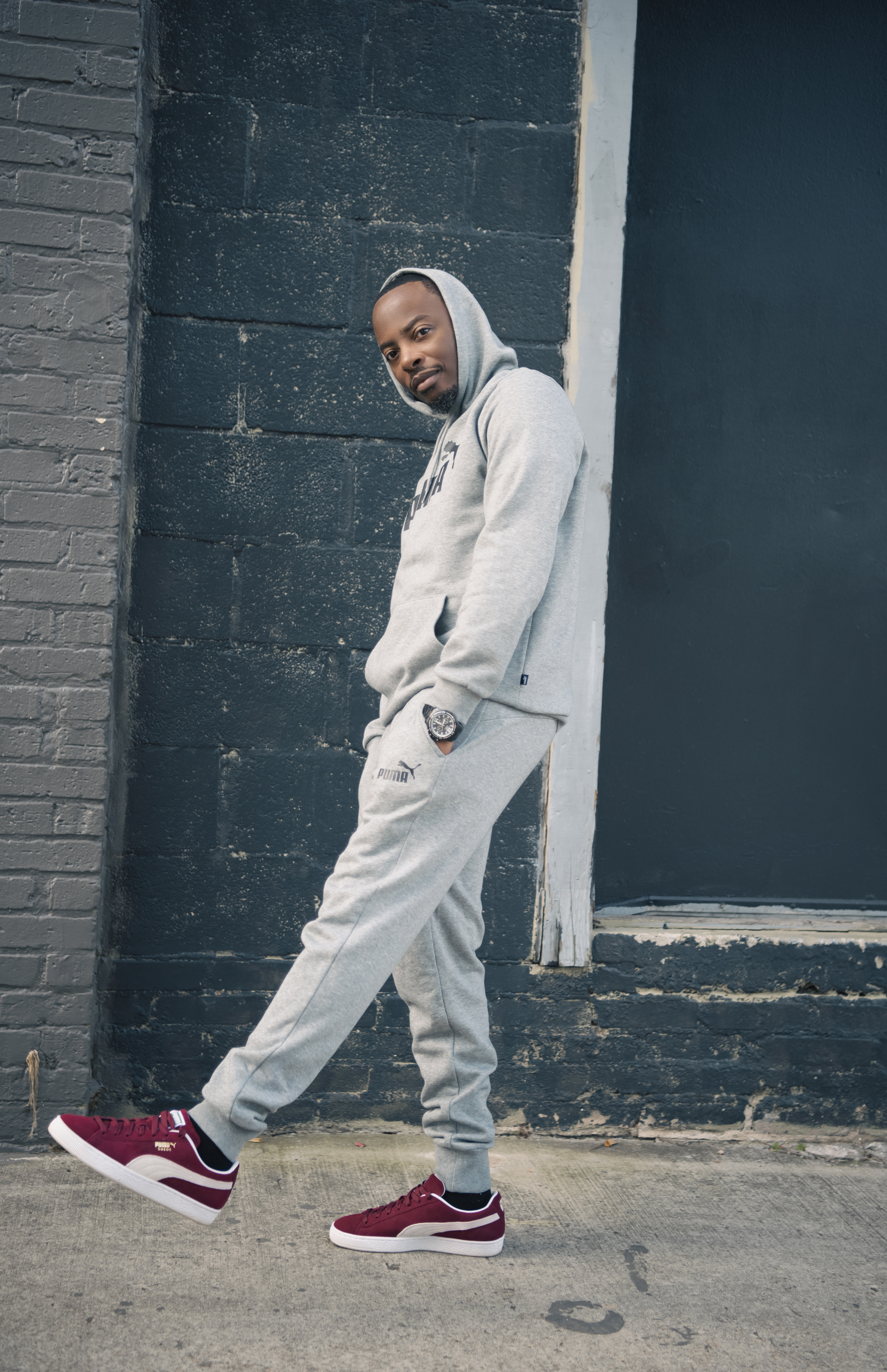 Walking pose in a gray Puma sweatsuit and burgundy suede sneakers against a dark brick wall