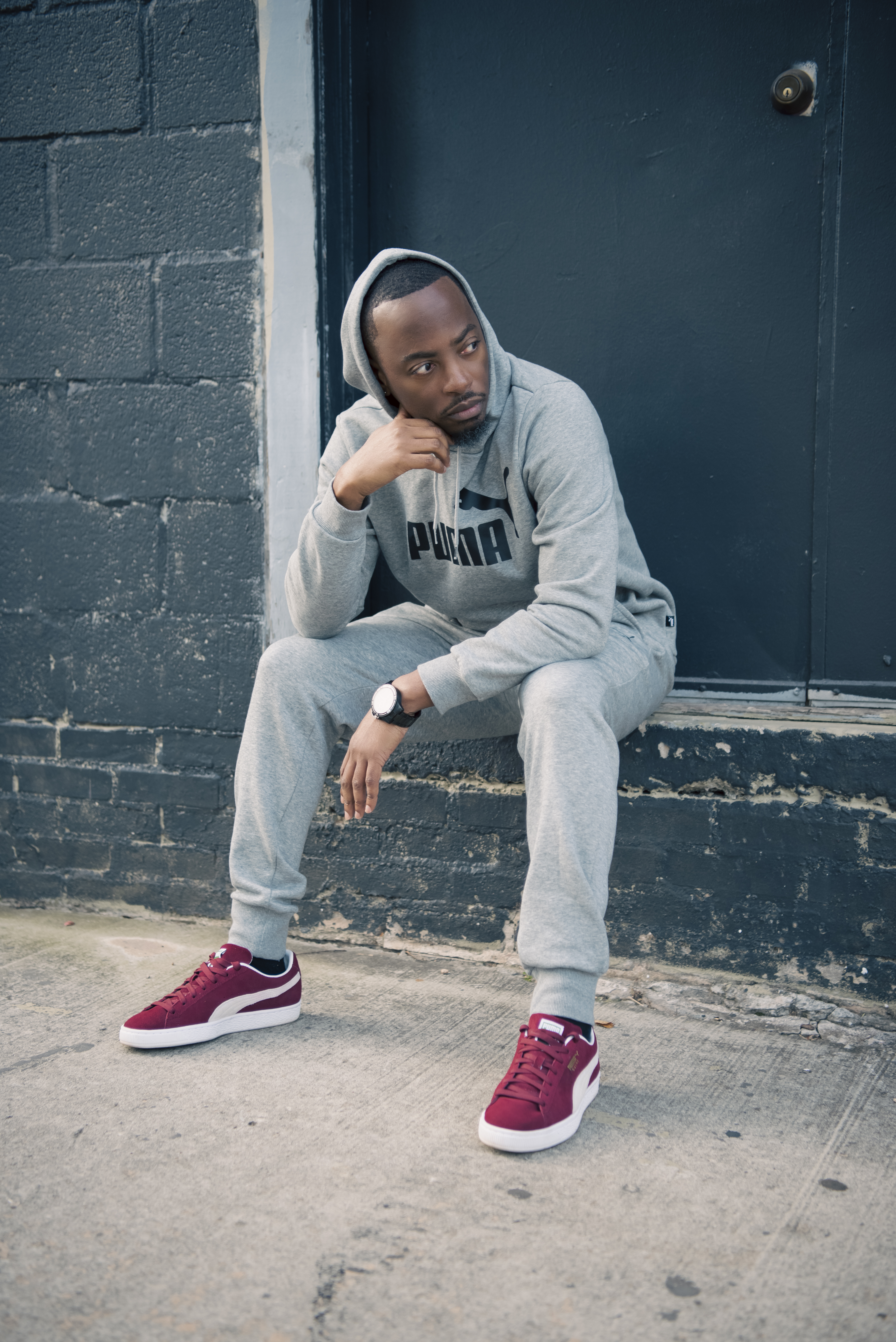 Seated on a step in a gray Puma sweatsuit and burgundy sneakers, hand to chin