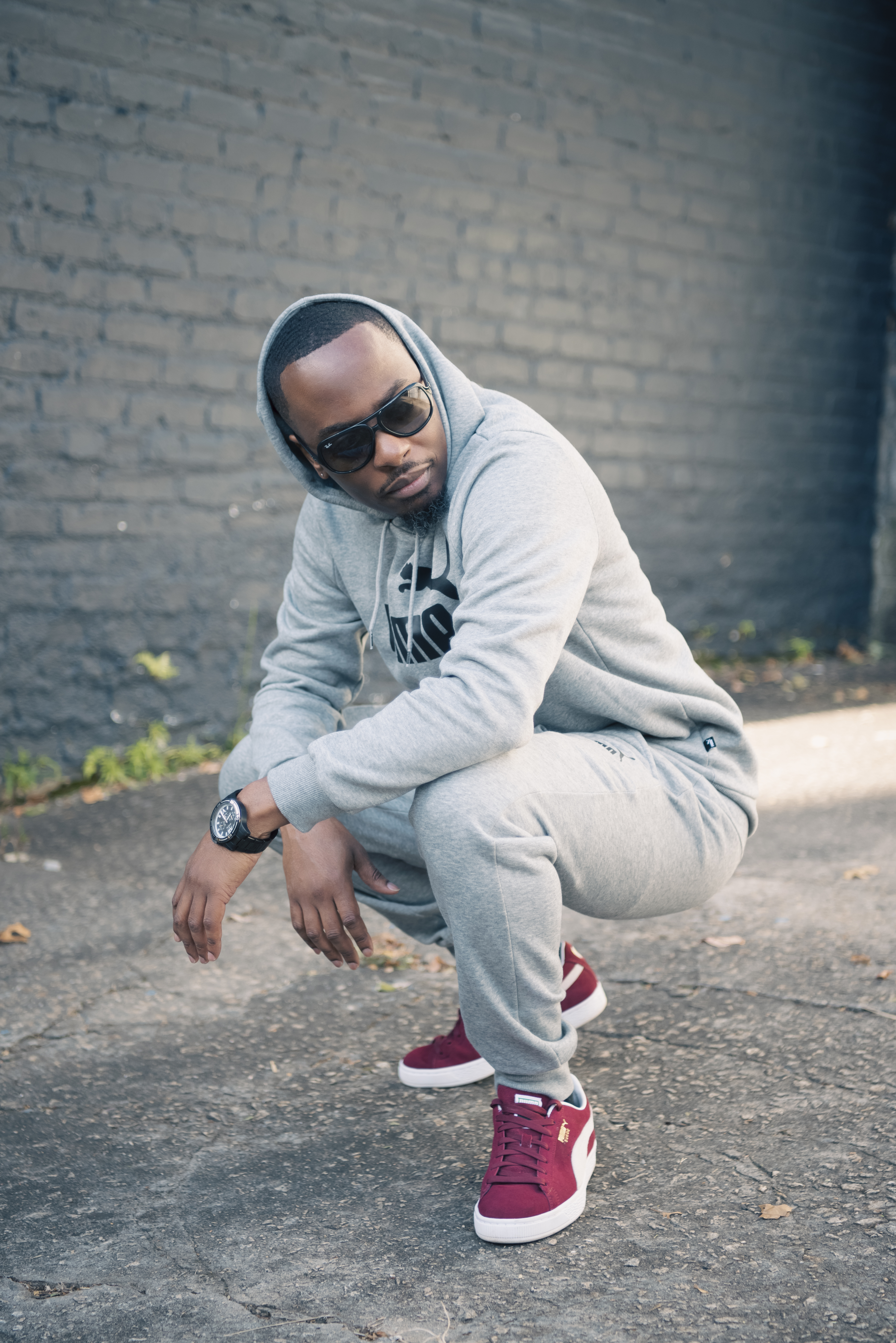Crouching in a gray Puma hoodie, sunglasses and burgundy sneakers